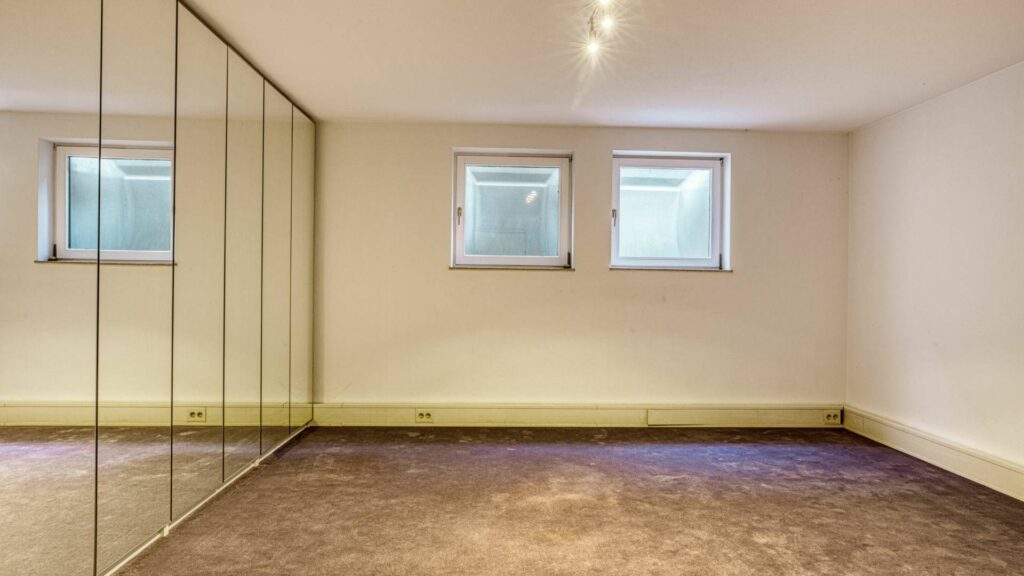 Unmöblierter Kellerraum mit Spiegelschrankfront und zwei kleinen Fenstern in einem Haus in München-Lerchenau