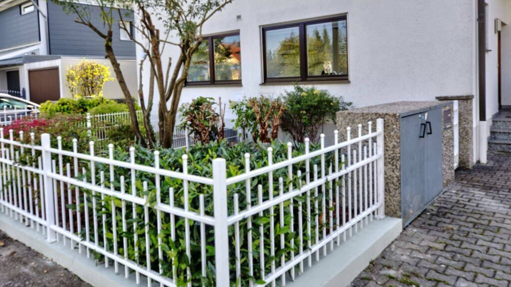 Gepflegter Vorgarten des Lerchenauer Hauses mit weißem Zaun, Hecken und Bepflanzung
