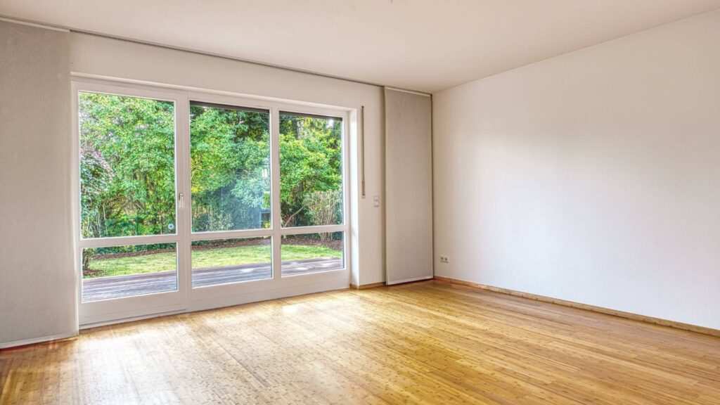 Leeres Wohnzimmer des Lerchenauer Hauses mit Parkettboden und breiter Fensterfront zum Garten