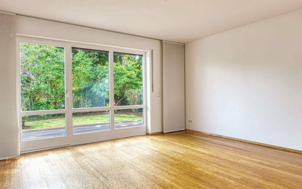 Leeres Wohnzimmer mit Parkettboden, bodentiefem Fenster zur Terrasse und Blick ins Grüne in Lerchenau.