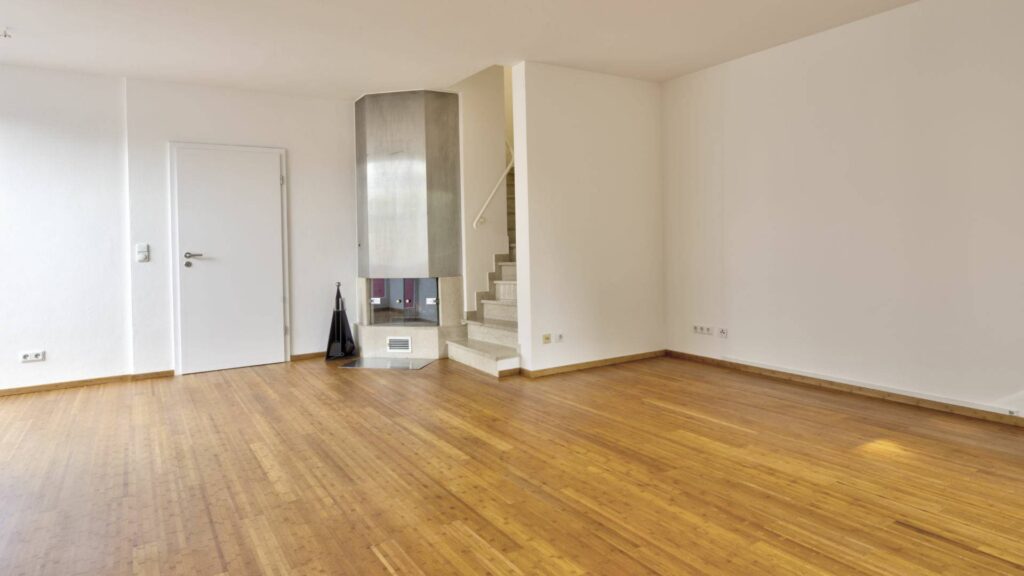 Leeres Wohnzimmer des Lerchenauer Hauses mit Parkettboden und freistehendem Kaminofen an der Treppe
