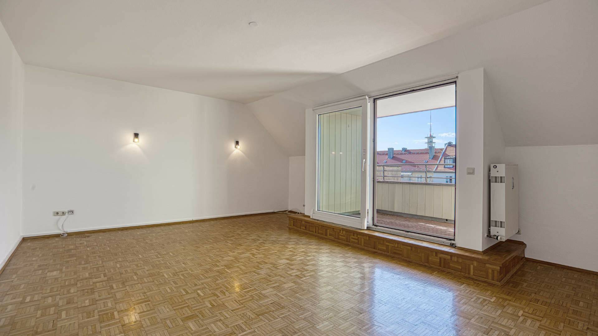 Leeres Wohnzimmer mit Dachschräge, Parkettboden und Balkontür mit Stadtblick in einer Dachgeschosswohnung in der Maxvorstadt
