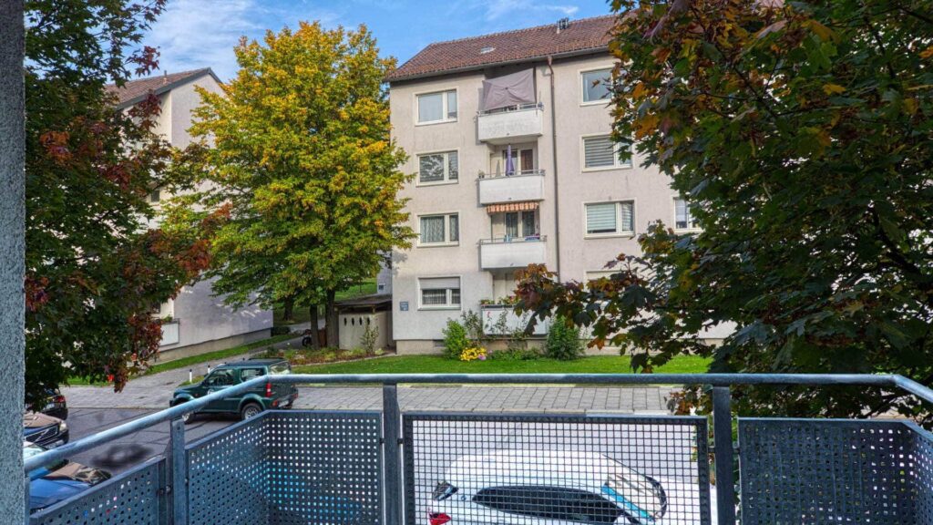 Blick vom Balkon auf ein Wohngebäude mit Herbstbäumen, parkenden Autos und einem Kettengeländer im Vordergrund.