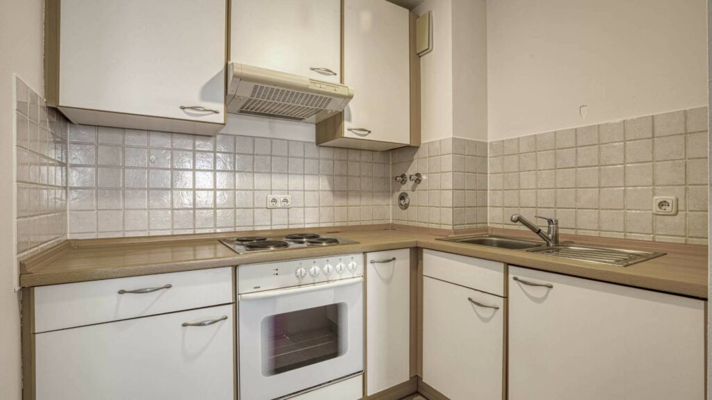 Kleine L-förmige Einbauküche mit weißen Fronten, Fliesenspiegel und freistehendem Herd in einer Wohnung in Moosach.