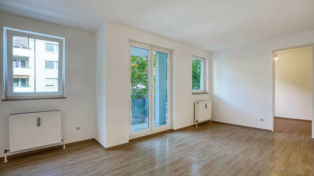 Leeres Wohnzimmer mit hellem Parkettboden, Fenster, Balkontür und Blick in einen weiteren Raum in Moosach.