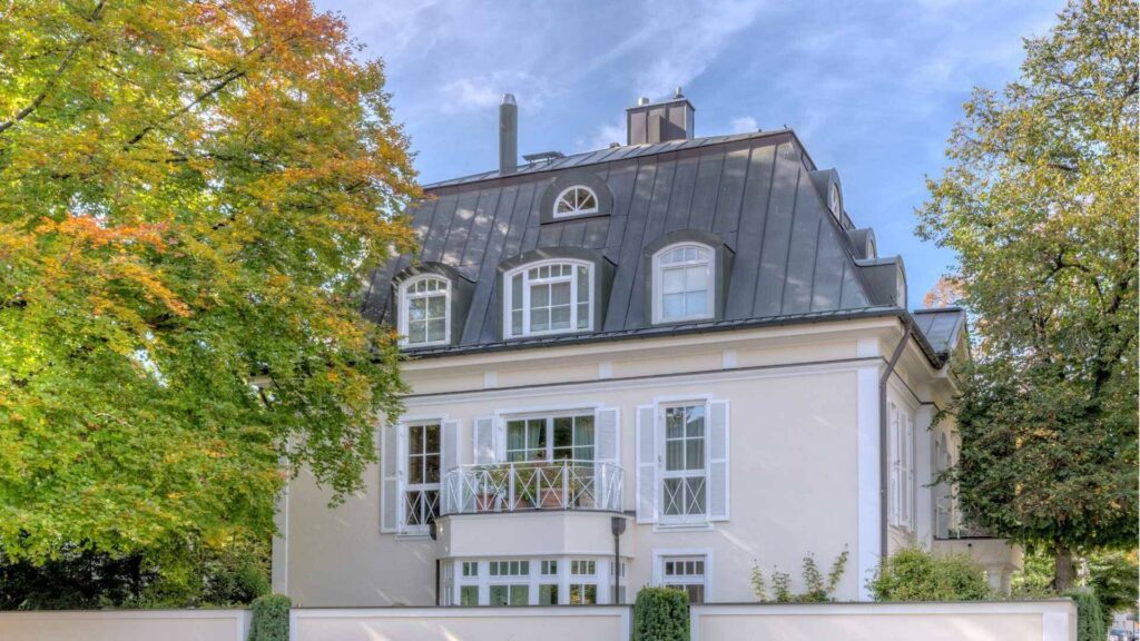 Weiße Villa mit dunklem Mansarddach umgeben von herbstlich gefärbten Bäumen