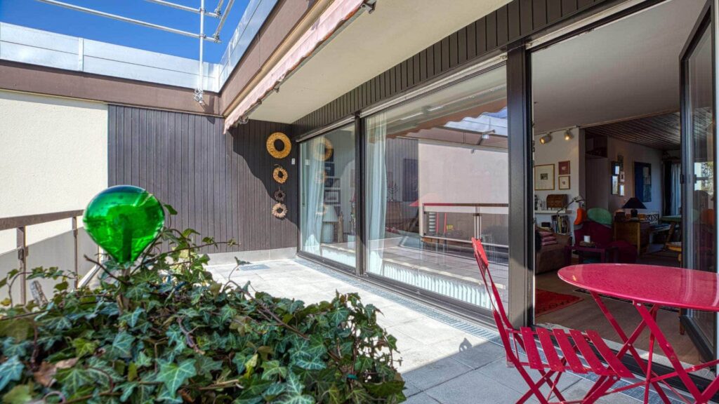 Gepflasterte Dachterrasse mit rotem Tisch, Stühlen und Blick durch die Glasfront ins Wohnzimmer in Obermenzing