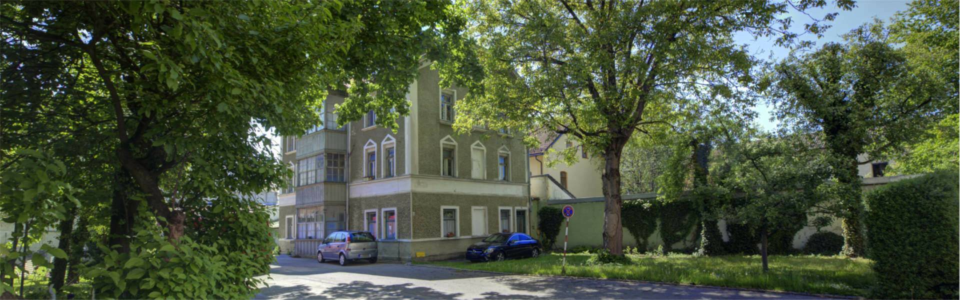 Historisches Gebäude mit gelber Fassade, umgeben von alten Bäumen und Hecken auf einem Grundstück in Rosenheim.