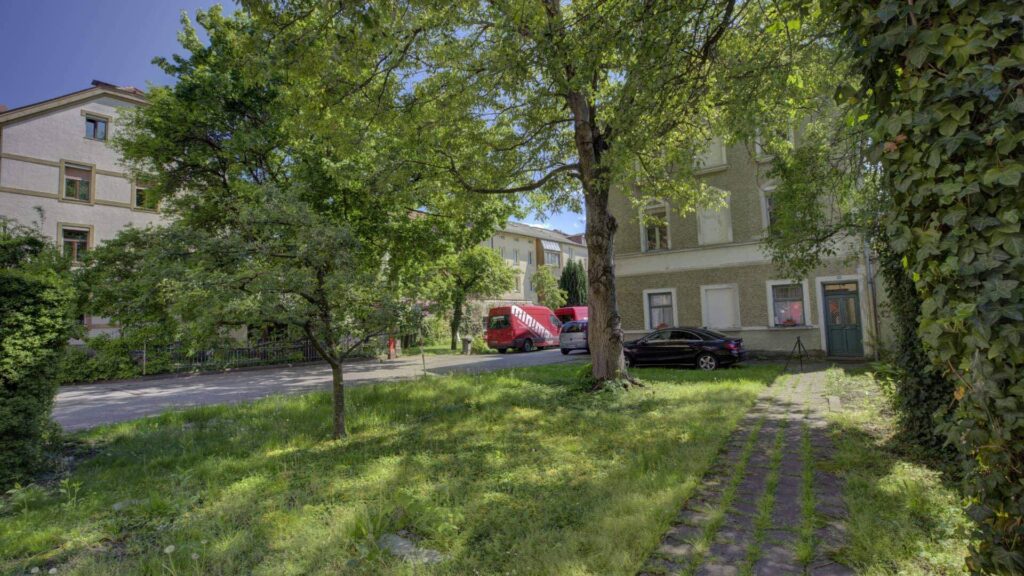 Grasbewachsene Zufahrt mit Bäumen und parkenden Autos vor einem Wohnhaus auf dem Grundstück in Rosenheim.