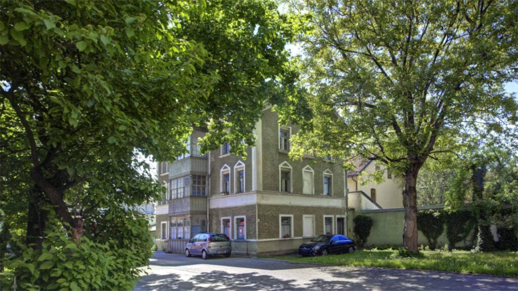 Straßenansicht eines historischen Gebäudes mit gelber Fassade, alten Bäumen und parkenden Autos in Rosenheim.