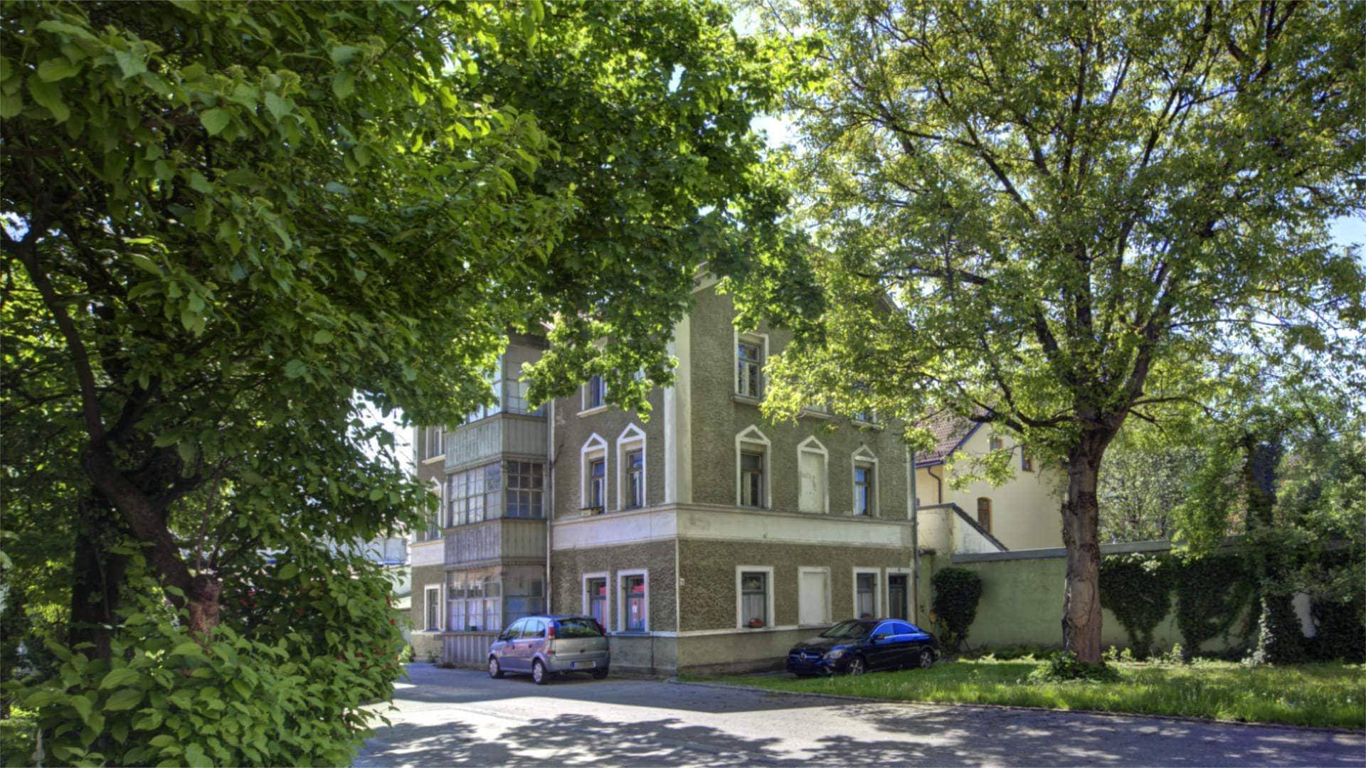 Straßenansicht eines historischen Gebäudes mit gelber Fassade, alten Bäumen und parkenden Autos in Rosenheim.