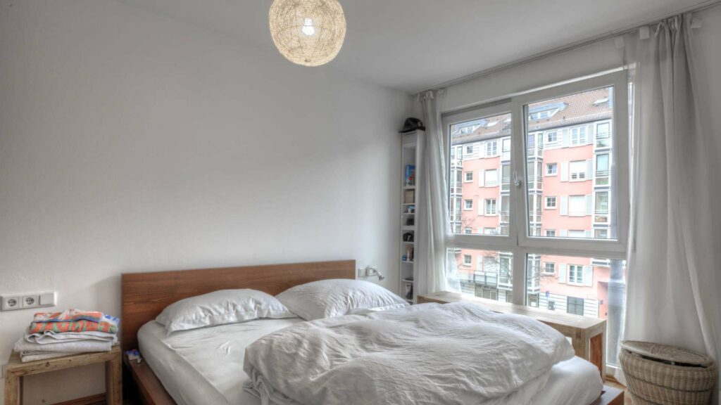 Schlafzimmer mit Doppelbett und Fensterblick auf Nachbargebäude in einer Wohnung in München-Sendling
