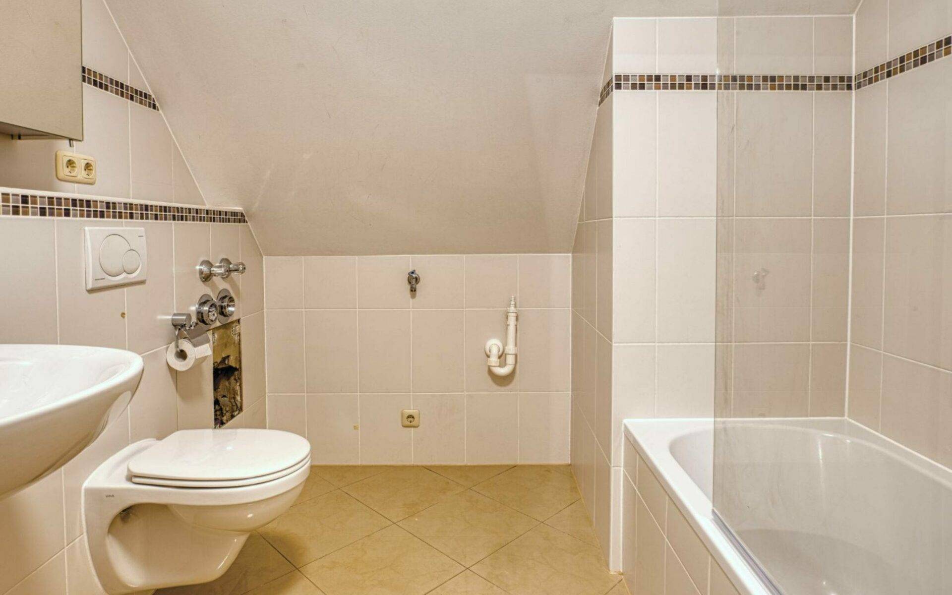 Badezimmer mit Wanne, WC, Waschbecken und dekorativem Fliesenband unter der Dachschräge in Thalkirchen.