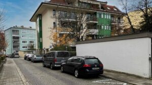 Straßenzug mit grün-weißem Wohnhaus, parkenden Autos und Bäumen in München-Thalkirchen.