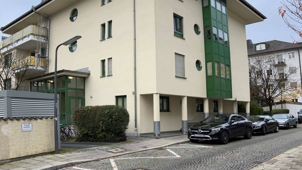 Cream-colored residential house with green window elements and parking spaces in front of the entrance in Munich-Thalkirchen.