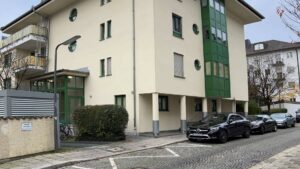 Cremefarbenes Wohnhaus mit grünen Fensterelementen und Parkplätzen vor dem Eingang in München-Thalkirchen.