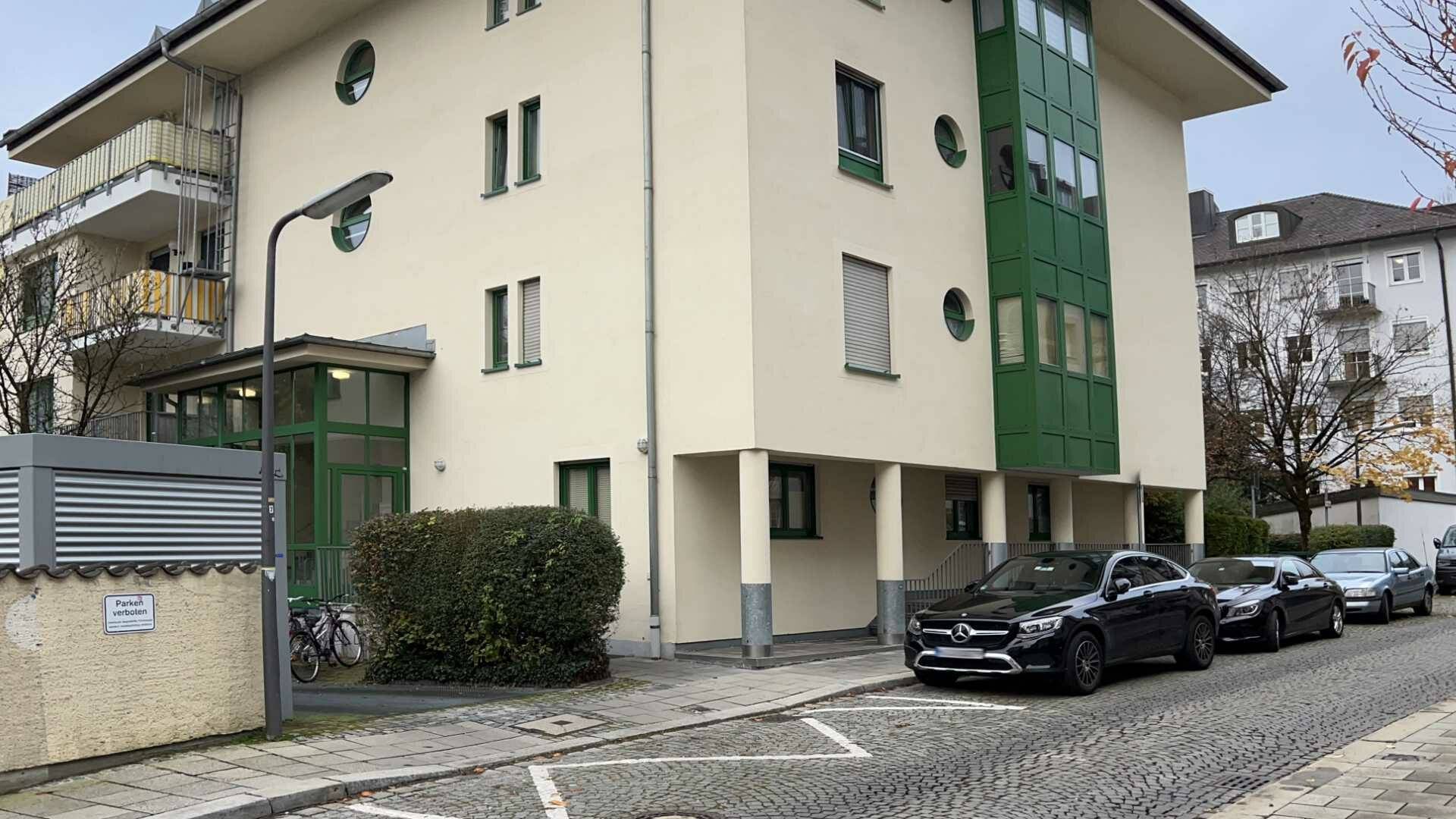 Cremefarbenes Wohnhaus mit grünen Fensterelementen und Parkplätzen vor dem Eingang in München-Thalkirchen.