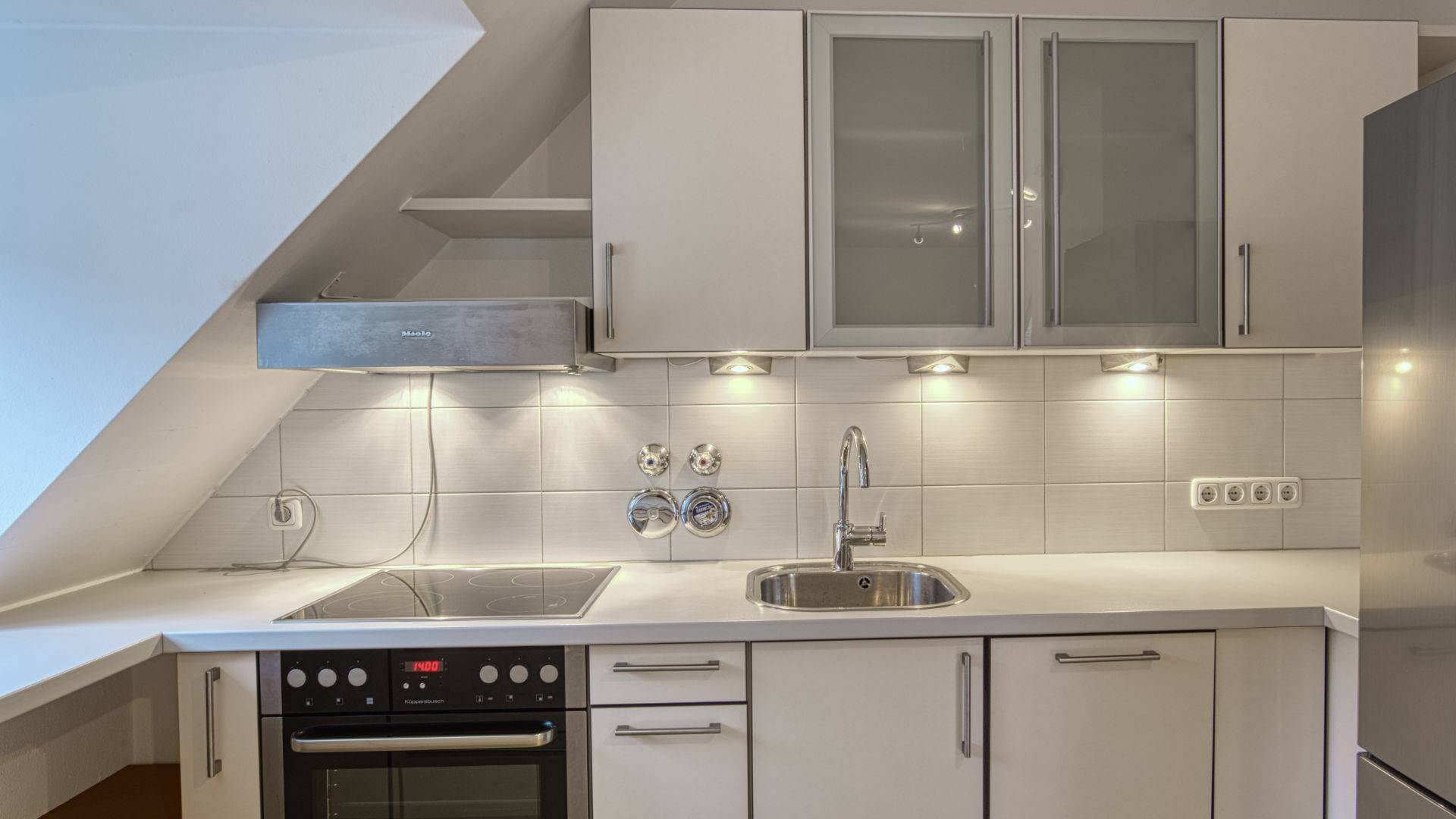 Weiße Einbauküche mit Glasfronten und Dachschräge in einer Dachgeschosswohnung in Thalkirchen.
