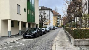 Ruhige Wohnstraße mit grün-weißem Eckhaus, parkenden Autos und Hecken in München-Thalkirchen.