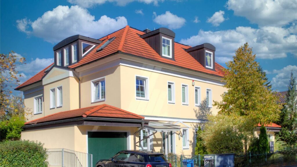 Gelbes Wohnhaus mit rotem Ziegeldach und Gauben an einer Straße