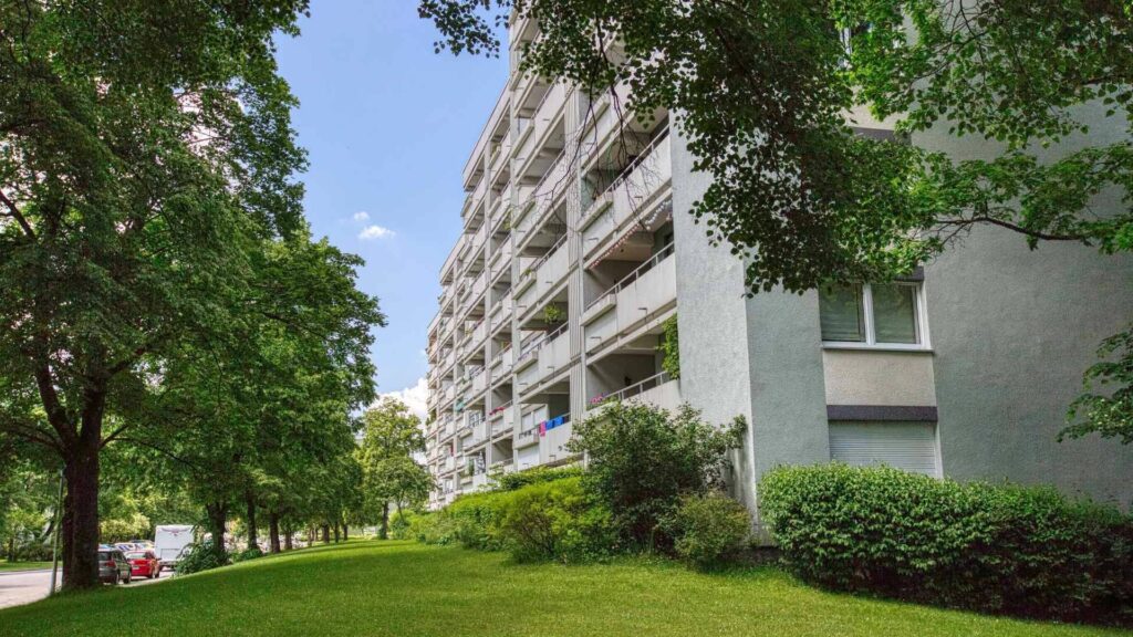 Außenansicht eines mehrstöckigen Wohnhauses mit Balkonen, umgeben von Bäumen und Rasenflächen in Unterhaching.