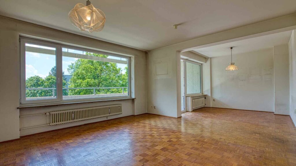 Leeres Wohnzimmer mit Parkettboden, großem Fenster mit Grünblick und Durchgang zu weiterem Raum in Unterhaching.