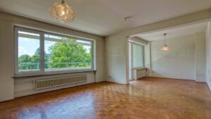 Leeres Wohnzimmer mit Parkettboden, großem Fenster mit Grünblick und Durchgang zu weiterem Raum in Unterhaching.