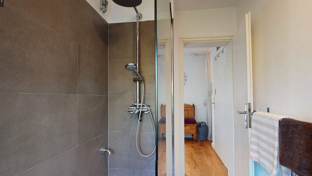 Walk-in shower with gray tiles and glass wall, view into the hallway of an apartment in Untermenzing.