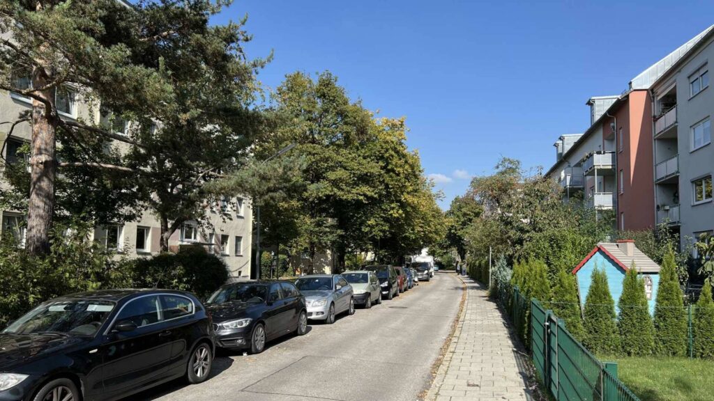 Tree-lined street with parked cars and a modern apartment building in Munich-Untermenzing.