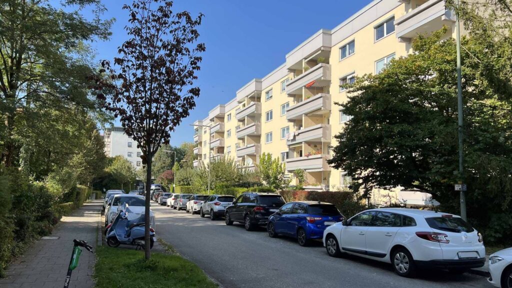 Residential street with a yellow apartment building, parked cars, and trees in Munich-Untermenzing.