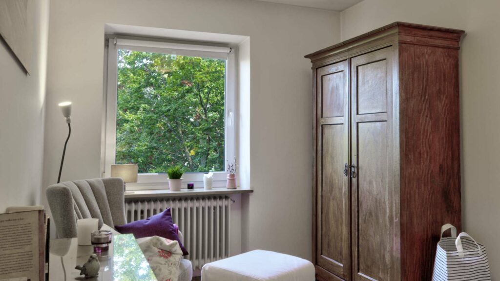 Inhabited room with an antique wooden wardrobe, sitting area, and window with a tree view in Untermenzing.
