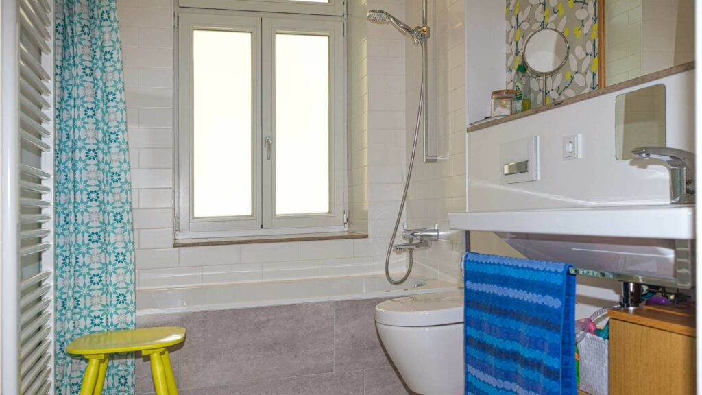 Badezimmer der Altbauwohnung in der Au mit Dusche, WC, Waschbecken und gelbem Hocker