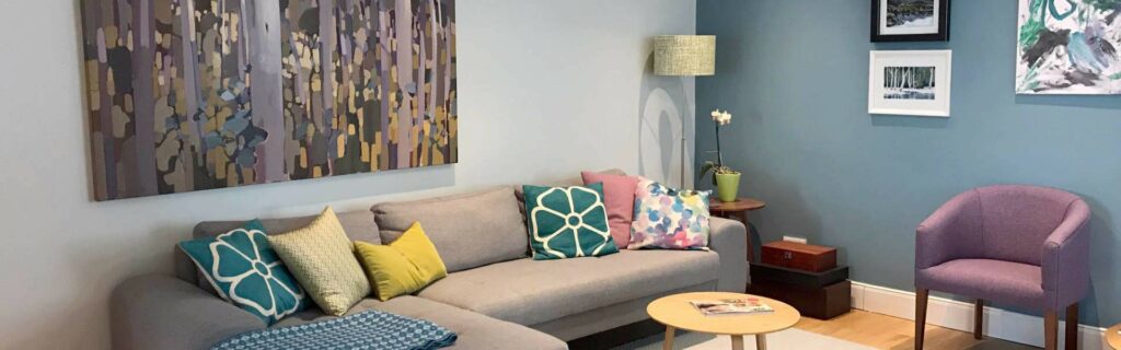 Wohnzimmer einer Altbauwohnung in der Au mit grauem Ecksofa und bunten Kissen sowie Kunstbildern.