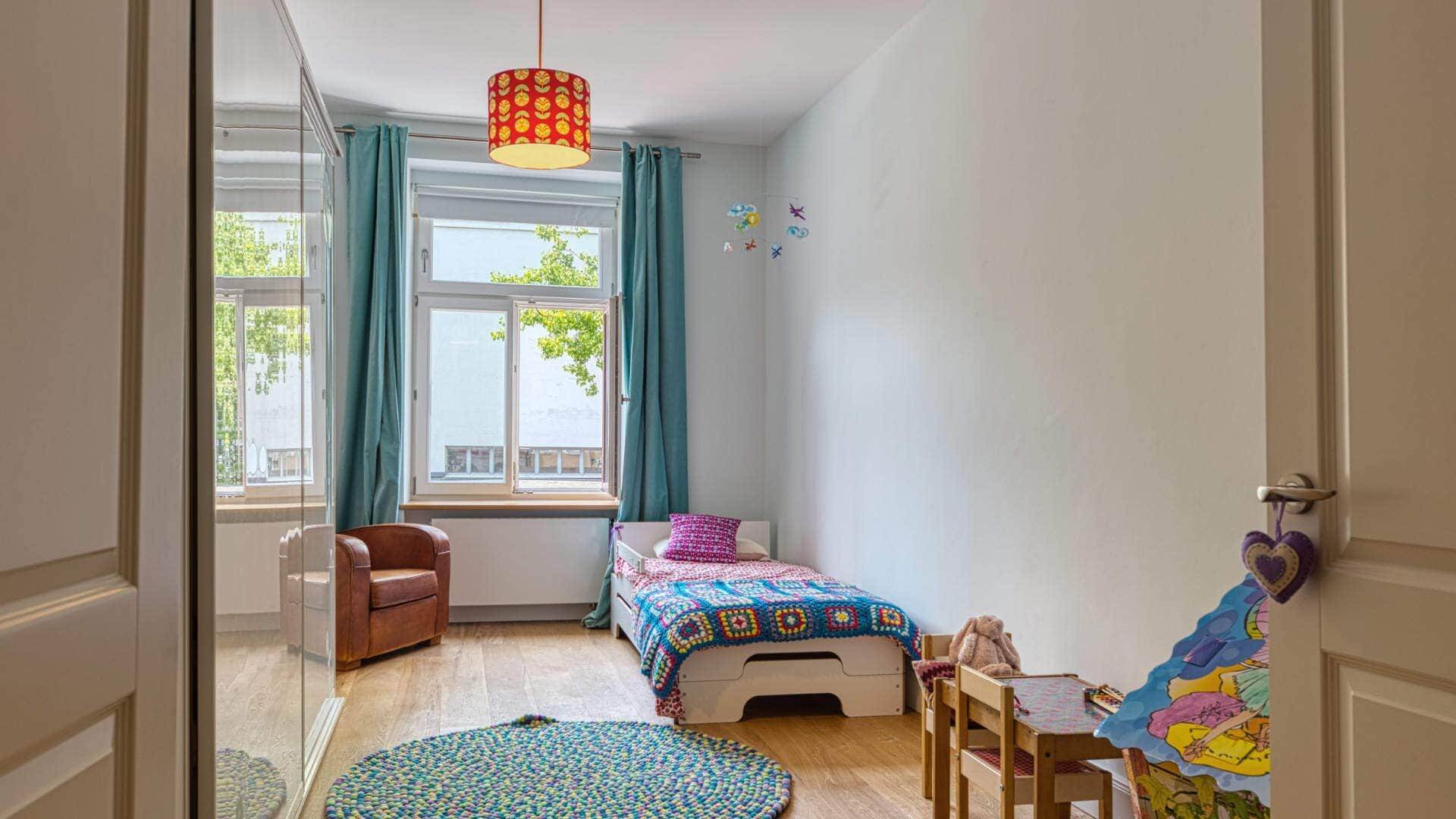 Children's room in an old-style apartment in Au with a bed, armchair, and high window with curtains.