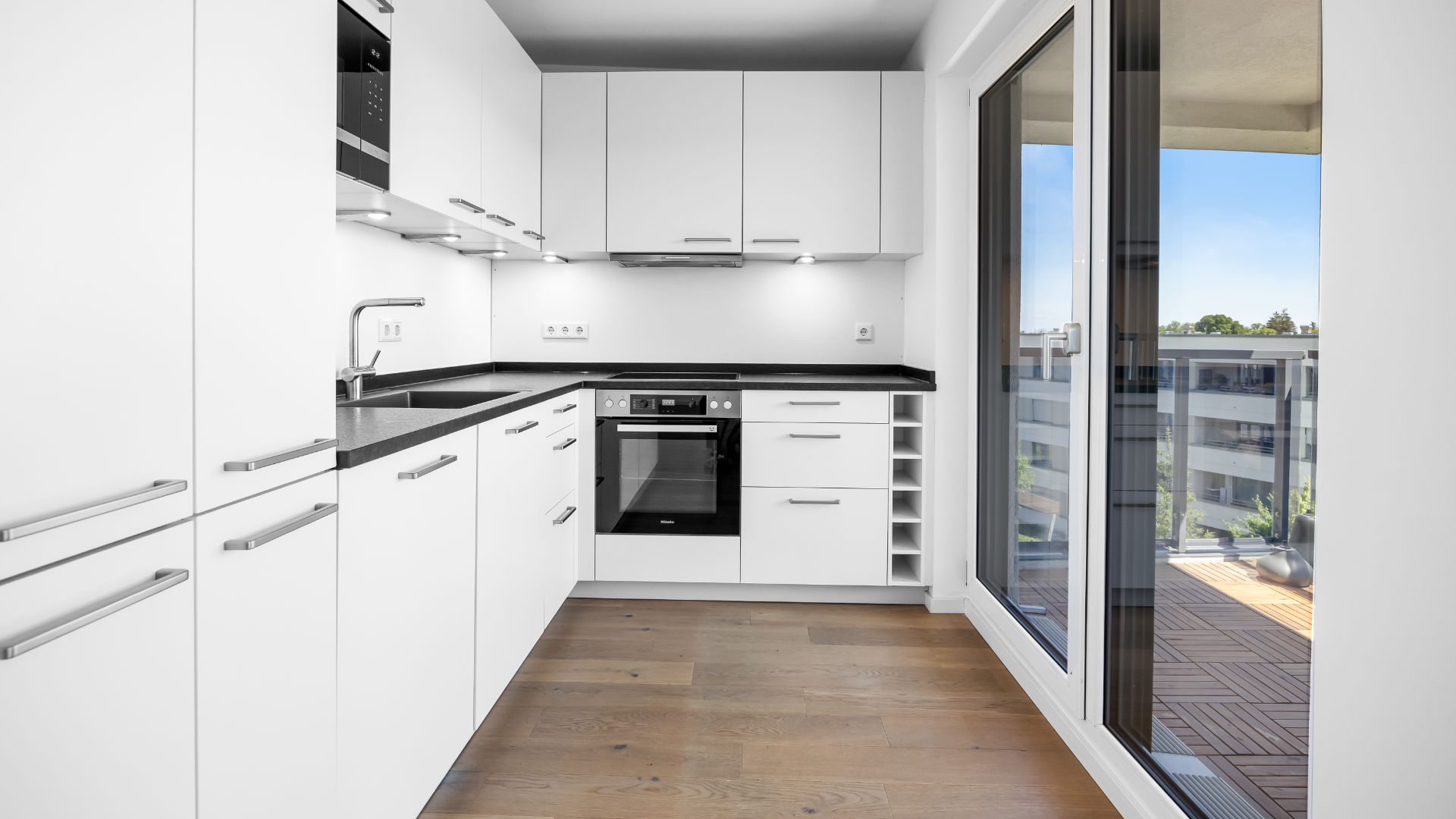 White kitchen of the new-build apartment in Aubing with a glass door to the wooden patio and a black countertop