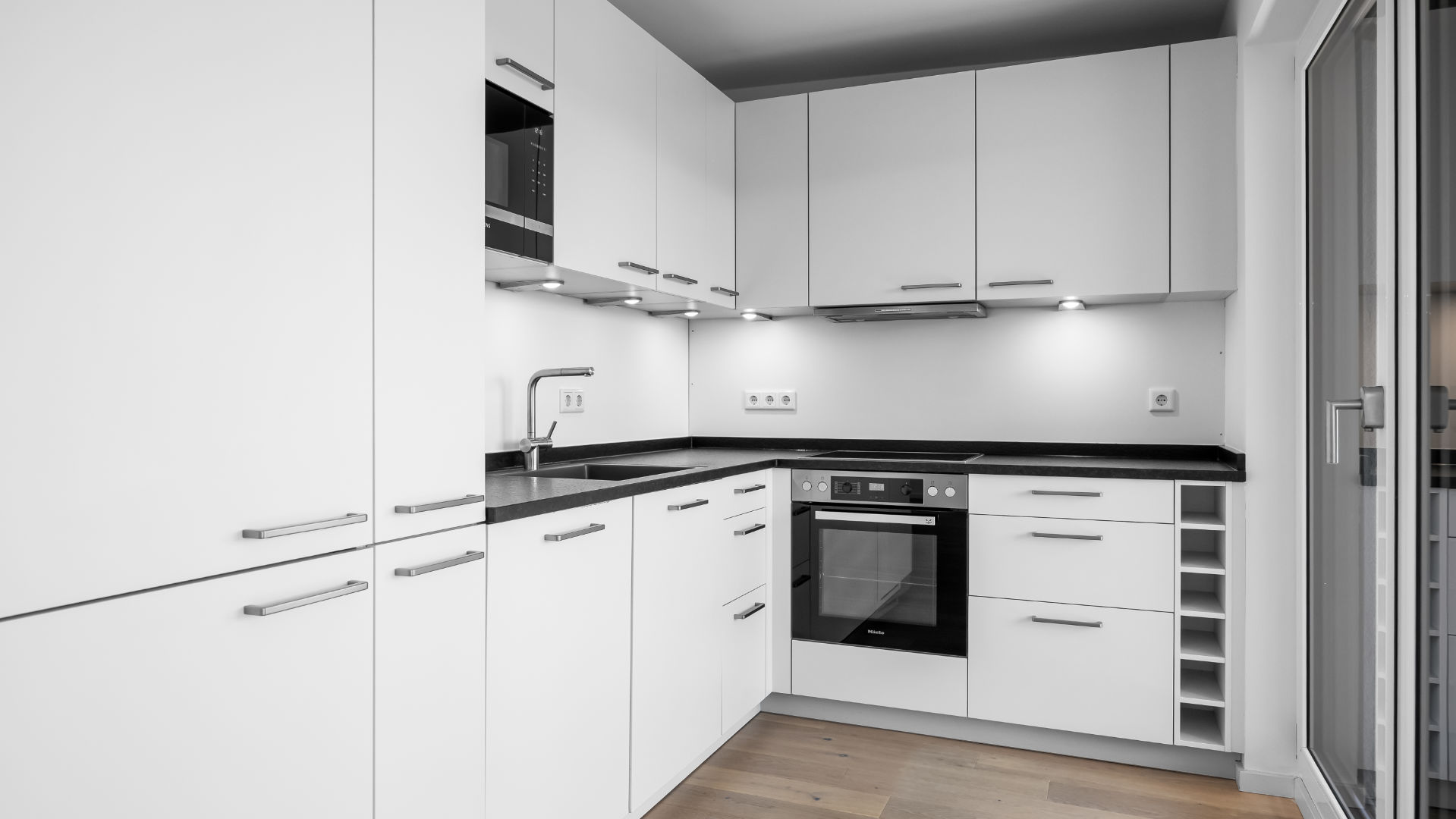 White fitted kitchen in the Aubing new-build apartment with a black countertop, oven, and wine rack