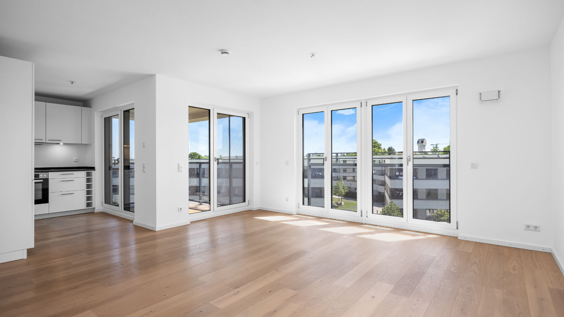 Empty living room with open kitchen and balcony access in the new-build apartment in Munich-Aubing – Rogers Immobilien