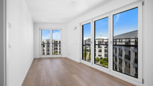 Empty room with a large floor-to-ceiling window and a view of the new Aubing residential development – Rogers Immobilien