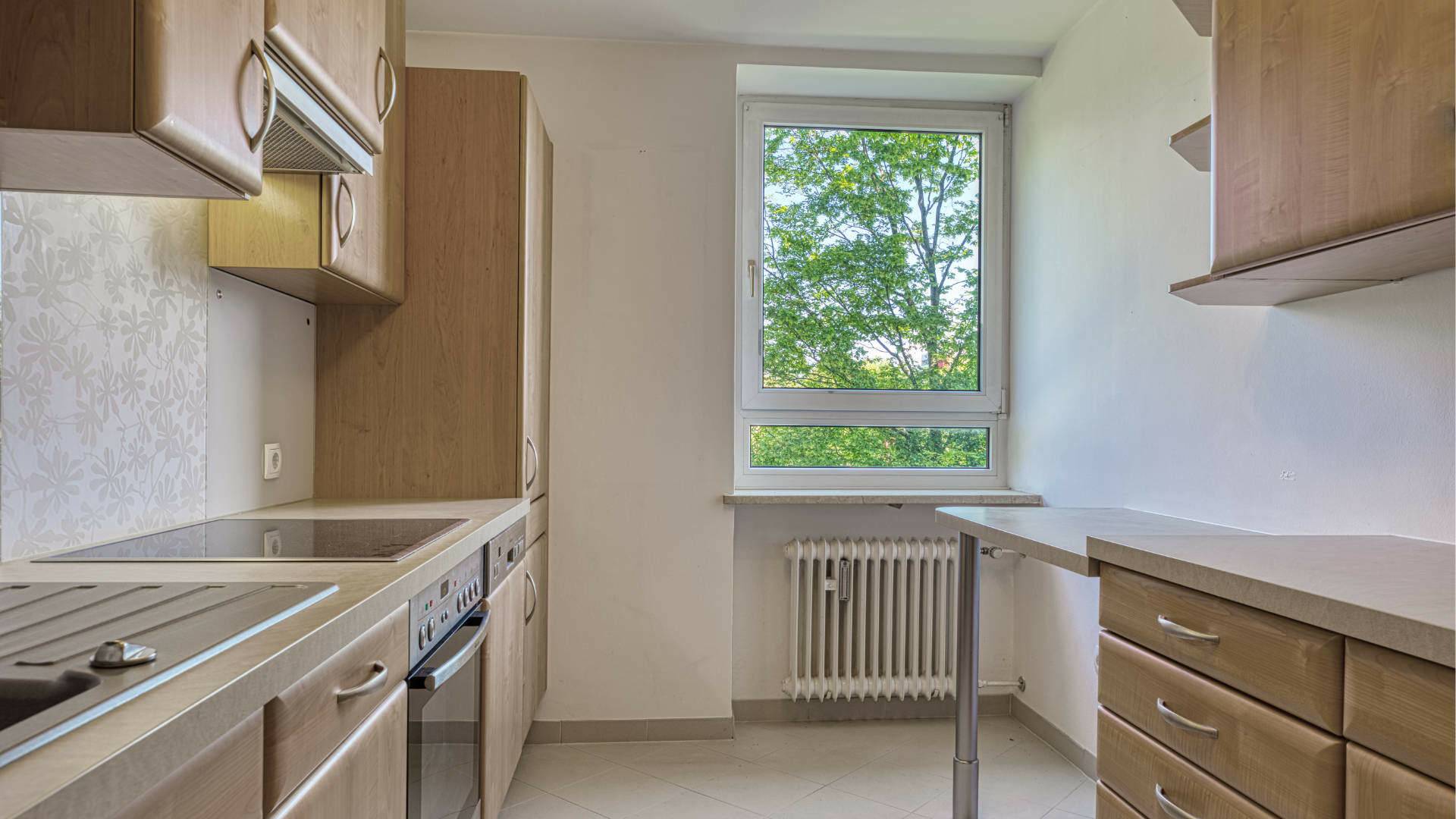 Kitchen in the apartment in Aubing with wooden fronts, sink, windows with views of the greenery, and heating pipes
