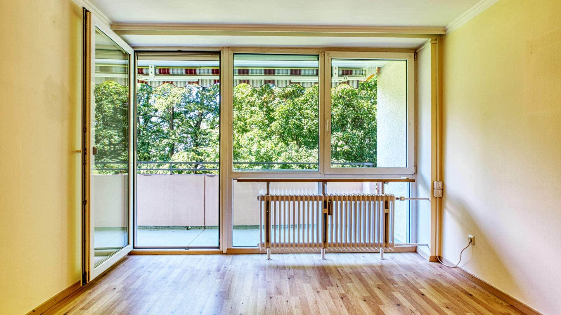 Empty living room in Aubing with a large window front, access to the balcony and light parquet floor.