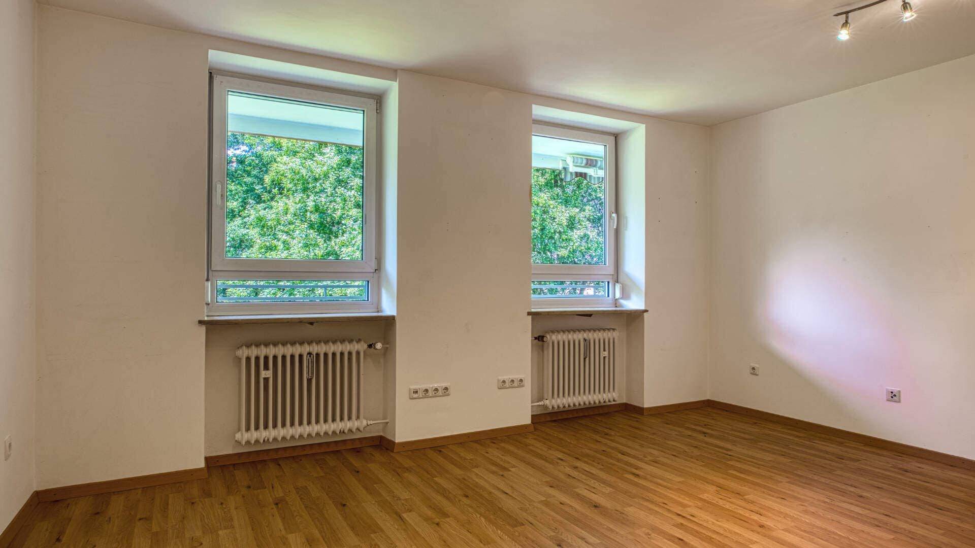 Empty room in the apartment in Aubing with two large windows, views of the greenery and parquet floor