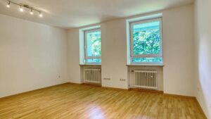 Empty room in Aubing with two windows, radiators and light parquet floor.