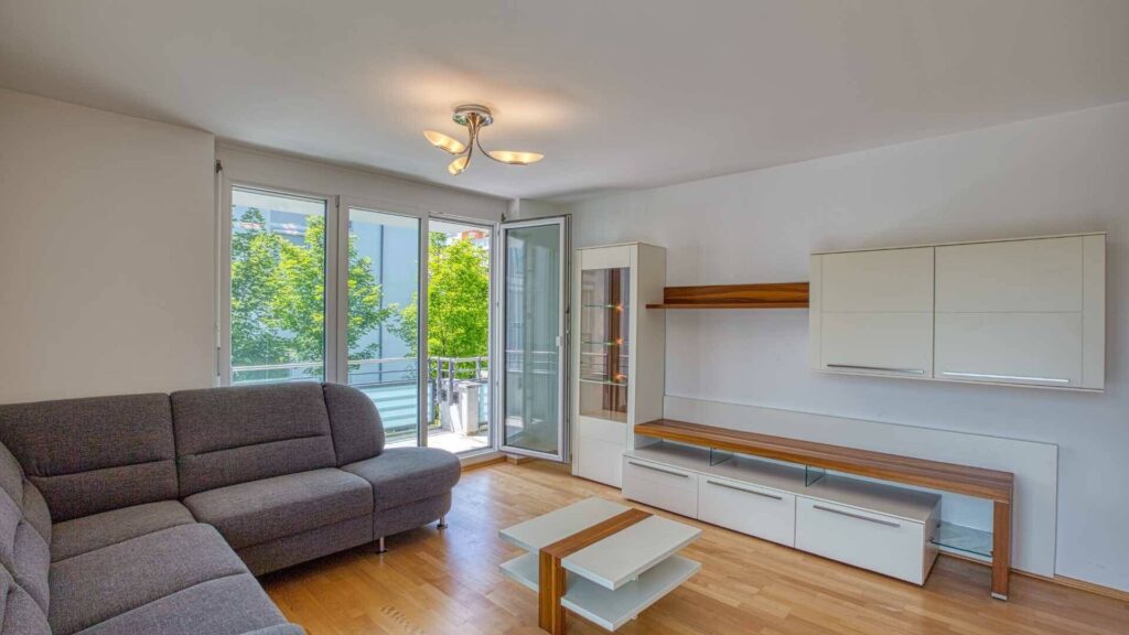Wohnzimmer in Bogenhausen mit grauem Sofa, Wohnwand und Balkonzugang.