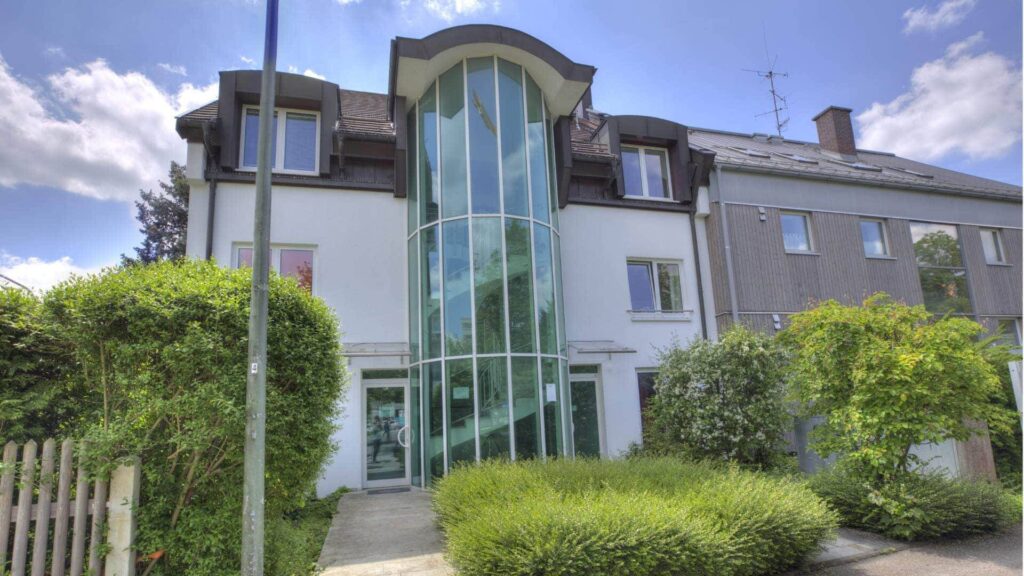 Weißes Gebäude mit markanter Glasfassade und Hecken, Bürostandort in München-Fürstenried.