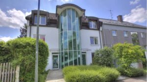 Weißes Gebäude mit markanter Glasfassade und Hecken, Bürostandort in München-Fürstenried.