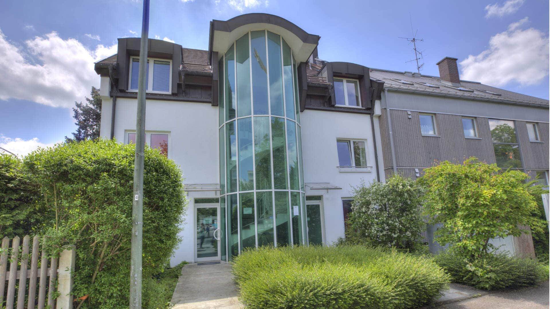 Weißes Gebäude mit markanter Glasfassade und Hecken, Bürostandort in München-Fürstenried.