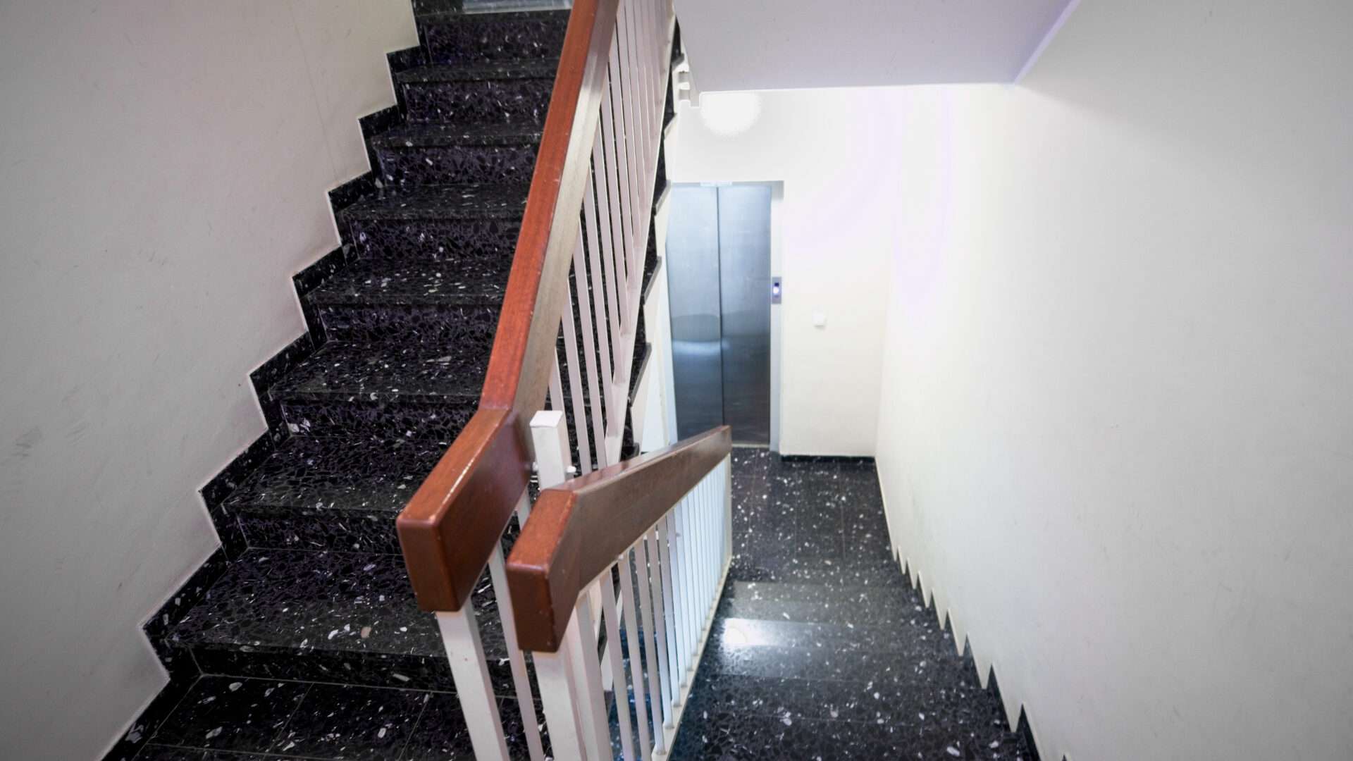 Treppenhaus mit dunklem Steinboden, Holzhandlauf und Wohnungstür in einem Mehrfamilienhaus in München-Giesing