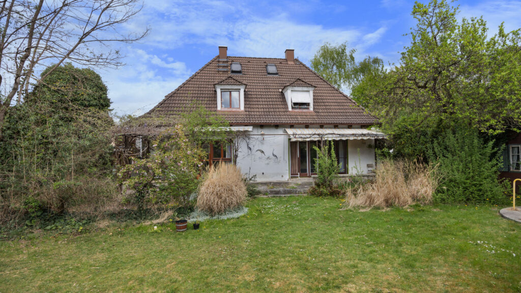Garden-side view of the existing house on the property in Großhadern with a hipped roof and overgrown lawn