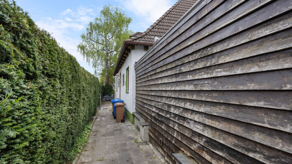 Narrow path between hedge and wooden facade of the existing building on the property in Großhadern – Rogers Immobilien