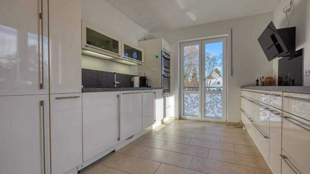 Moderne weiße Hochglanzküche in Hadern mit dunkler Arbeitsplatte und Terrassenzugang.